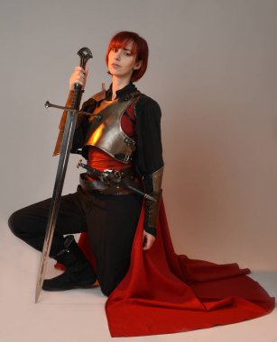 Fantezi savaşçısı kadın modelinin portresi, elinde kılıç, kırmızı peri masalı pelerinli tarihi ortaçağ zırhı, güçlü bir fantezi şövalyesi. Stüdyonun arka planında izole bir şekilde oturuyor.