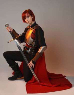 Fantezi savaşçısı kadın modelinin portresi, elinde kılıç, kırmızı peri masalı pelerinli tarihi ortaçağ zırhı, güçlü bir fantezi şövalyesi. Stüdyonun arka planında izole bir şekilde oturuyor.