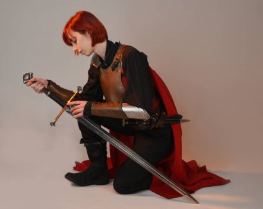 Fantezi savaşçısı kadın modelinin portresi, elinde kılıç, kırmızı peri masalı pelerinli tarihi ortaçağ zırhı, güçlü bir fantezi şövalyesi. Stüdyonun arka planında izole bir şekilde oturuyor.