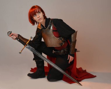 Fantezi savaşçısı kadın modelinin portresi, elinde kılıç, kırmızı peri masalı pelerinli tarihi ortaçağ zırhı, güçlü bir fantezi şövalyesi. Stüdyonun arka planında izole bir şekilde oturuyor.