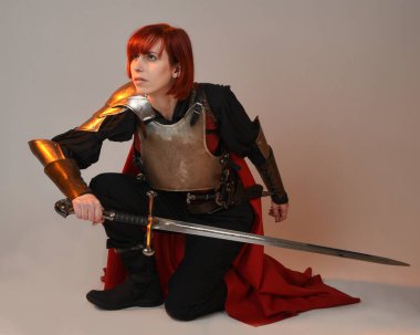 Fantezi savaşçısı kadın modelinin portresi, elinde kılıç, kırmızı peri masalı pelerinli tarihi ortaçağ zırhı, güçlü bir fantezi şövalyesi. Stüdyonun arka planında izole bir şekilde oturuyor.