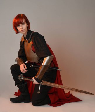 Fantezi savaşçısı kadın modelinin portresi, elinde kılıç, kırmızı peri masalı pelerinli tarihi ortaçağ zırhı, güçlü bir fantezi şövalyesi. Stüdyonun arka planında izole bir şekilde oturuyor.