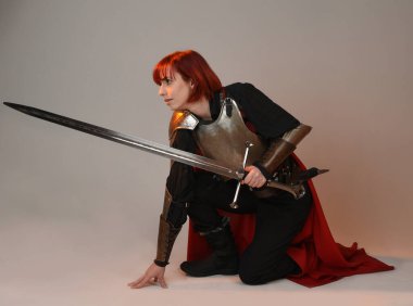 Fantezi savaşçısı kadın modelinin portresi, elinde kılıç, kırmızı peri masalı pelerinli tarihi ortaçağ zırhı, güçlü bir fantezi şövalyesi. Stüdyonun arka planında izole bir şekilde oturuyor.