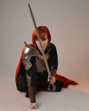 Fantezi savaşçısı kadın modelinin portresi, elinde kılıç, kırmızı peri masalı pelerinli tarihi ortaçağ zırhı, güçlü bir fantezi şövalyesi. Stüdyonun arka planında izole bir şekilde oturuyor.