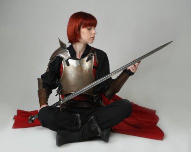 Fantezi savaşçısı kadın modelinin portresi, elinde kılıç, kırmızı peri masalı pelerinli tarihi ortaçağ zırhı, güçlü bir fantezi şövalyesi. Stüdyonun arka planında izole bir şekilde oturuyor.