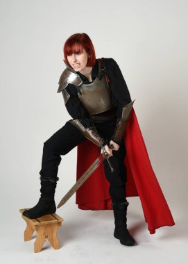 Tarihsel ortaçağ zırhlı fantezi savaşçı kadın modeli, kırmızı peri masalı pelerini, güçlü bir fantezi şövalyesinin kılıç tutan sinematik portresi. Epic standing poz, tecrit edilmiş beyaz stüdyo arka planı