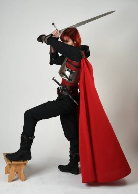 Fantezi savaşçısı kadın model, tarihi ortaçağ zırhı ve kırmızı peri masalı pelerini içinde kızıl saçlı güçlü bir fantezi şövalyesinin sinematik portresi, stüdyo arka planında dramatik bir duruş.