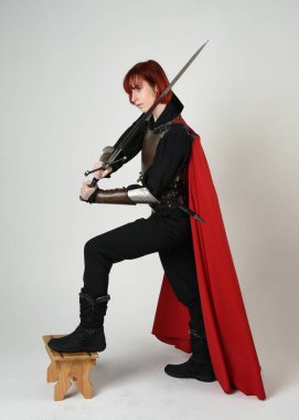 Fantezi savaşçısı kadın model, tarihi ortaçağ zırhı ve kırmızı peri masalı pelerini içinde kızıl saçlı güçlü bir fantezi şövalyesinin sinematik portresi, stüdyo arka planında dramatik bir duruş.