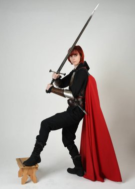 Fantezi savaşçısı kadın model, tarihi ortaçağ zırhı ve kırmızı peri masalı pelerini içinde kızıl saçlı güçlü bir fantezi şövalyesinin sinematik portresi, stüdyo arka planında dramatik bir duruş.