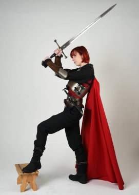 Fantezi savaşçısı kadın model, tarihi ortaçağ zırhı ve kırmızı peri masalı pelerini içinde kızıl saçlı güçlü bir fantezi şövalyesinin sinematik portresi, stüdyo arka planında dramatik bir duruş.