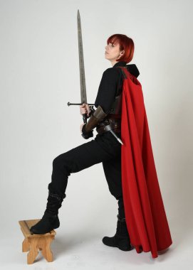 Fantezi savaşçısı kadın model, tarihi ortaçağ zırhı ve kırmızı peri masalı pelerini içinde kızıl saçlı güçlü bir fantezi şövalyesinin sinematik portresi, stüdyo arka planında dramatik bir duruş.