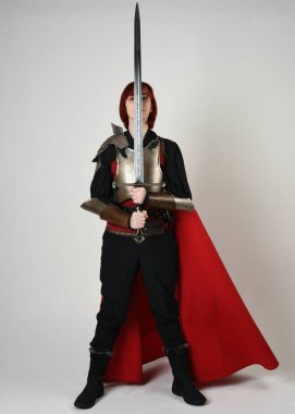 Tarihsel ortaçağ zırhlı fantezi savaşçı kadın modeli, kırmızı peri masalı pelerini, güçlü bir fantezi şövalyesinin kılıç tutan sinematik portresi. Epic standing poz, tecrit edilmiş beyaz stüdyo arka planı