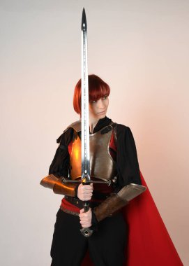 Fantezi savaşçısı kadın modelinin yakın plan portresi, tarihi ortaçağ zırh kostümü, kırmızı peri masalı pelerini, güçlü bir fantezi şövalyesi, kılıç taşıyor. izole edilmiş stüdyo arka planı altın sinematik ışıklandırma.