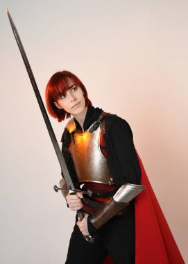 Fantezi savaşçısı kadın modelinin yakın plan portresi, tarihi ortaçağ zırh kostümü, kırmızı peri masalı pelerini, güçlü bir fantezi şövalyesi, kılıç taşıyor. izole edilmiş stüdyo arka planı altın sinematik ışıklandırma.