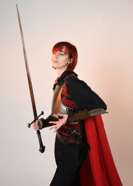 Fantezi savaşçısı kadın modelinin yakın plan portresi, tarihi ortaçağ zırh kostümü, kırmızı peri masalı pelerini, güçlü bir fantezi şövalyesi, kılıç taşıyor. izole edilmiş stüdyo arka planı altın sinematik ışıklandırma.