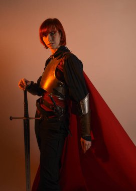 Fantezi savaşçısı kadın modelinin portresi, elinde kılıç, kırmızı peri masalı pelerinli tarihi ortaçağ zırhı, güçlü bir fantezi şövalyesi. izole edilmiş stüdyo arka planı altın sinematik ışıklandırma.