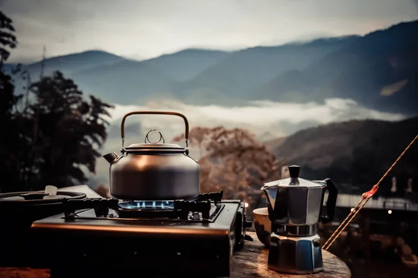 Outdoor kitchen equipment camp fire and brewing tea pot moka coffee drip cup,with wooden table camping gas stove set in nature outdoor,mountain background,concept camp and travel,vintage tone color