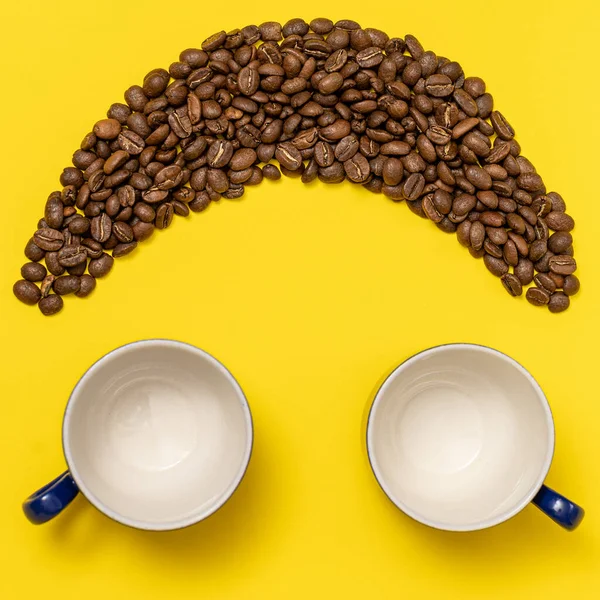 Happy emoticon made of coffee beans on a yellow background, Smiley made of coffee and two empty cups as eyes.