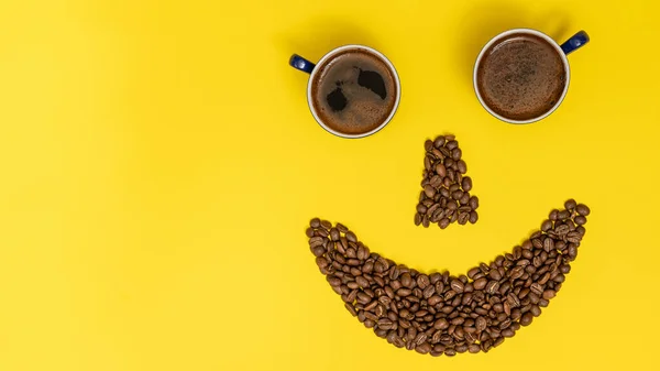 Smiling emoticon, lips made of coffee beans and eyes made of coffee cups, composition on a yellow background.