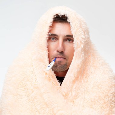 A sick man wrapped in a blanket holds a thermometer in his mouth, healthcare and treatment.