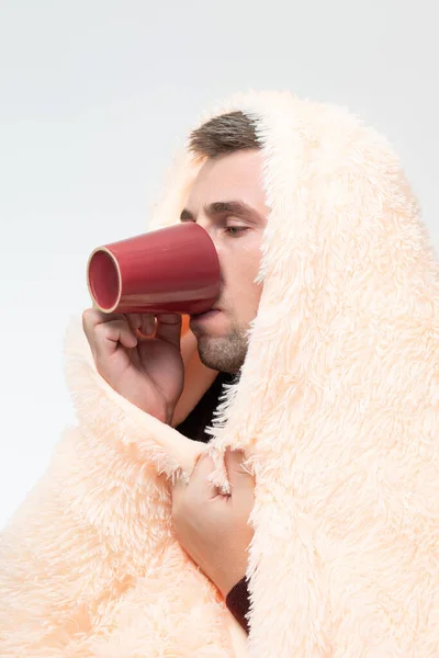 A man is wrapped in a blanket, holding a mug with a drink in his hands, a man is sick with the flu, a portrait of a sick man.