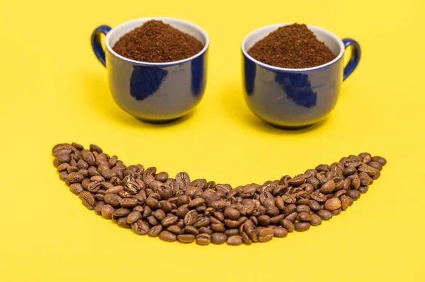 Happy emoticon made of coffee beans on a yellow background, Smiley made of coffee and two cups of ground coffee as eyes.