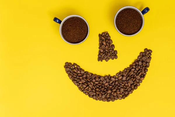 Smiling emoticon, lips made of coffee beans and eyes made of coffee cups, composition on a yellow background.