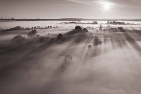 Flying over the village at dawn, bright sun on the horizon and fog over a small village, thick clouds and fog and clear sky, black and white photo.