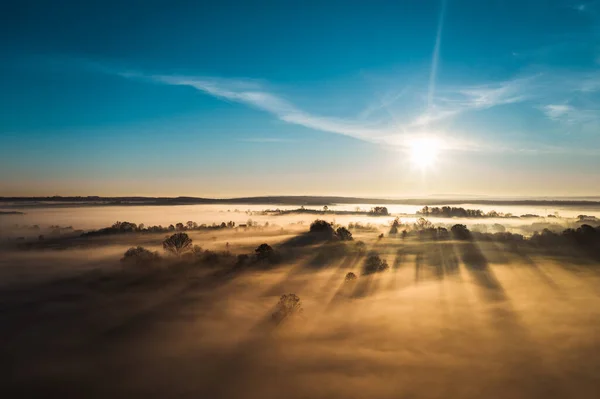 Flying over a village at dawn, bright sun on the horizon and fog over a small village, thick clouds and fog and a clear sky.