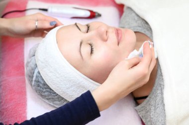 A woman cosmetologist washes a client with cotton wool and water, the client is wearing a disposable cap and is lying on the couch.