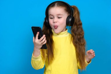 Portrait of a little girl with a phone and wireless headphones, a child in a yellow sweater on a blue background, a child shows his tongue.