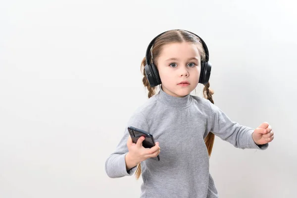 Happy little girl with wireless headphones and phone, portrait of girl with pigtails on white background with headphones and smartphone, photo with copy space.