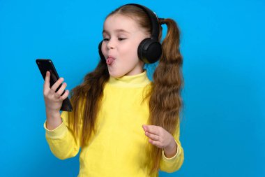 Portrait of a little girl with a phone and wireless headphones, a child in a yellow sweater on a blue background, a child shows his tongue.