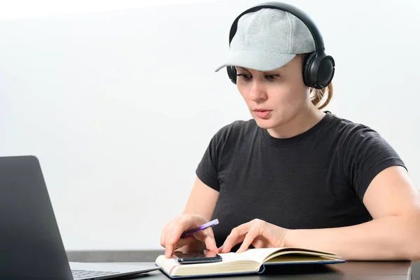 Busy young woman in headphones working on laptop, making video call and making notes in notebook, work and study online, copy space and white background.