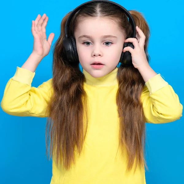 Happy little girl with wireless headphones on blue background doing dance moves to music in headphones, child with long hair.