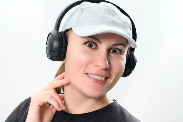 在白色背景上的女孩的肖像 头戴一顶帽子 头戴无线耳机 带着耳机的女孩的情感照片 — 图库照片