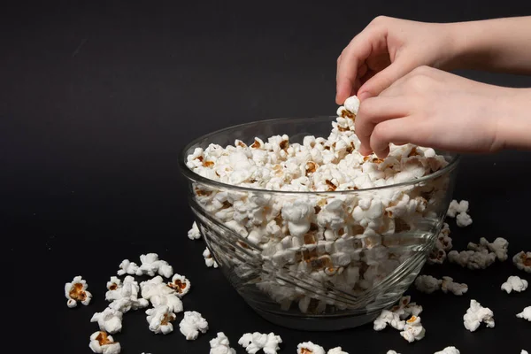 A child takes popcorn from a bowl, scattered popcorn near a plate, black background and popcorn, copy space and close-up.
