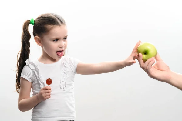 小さな女の子は リンゴ お菓子や果物の上にロリポップを選択し 健康的で不健康な食べ物 白い背景と子供の間の選択 — ストック写真
