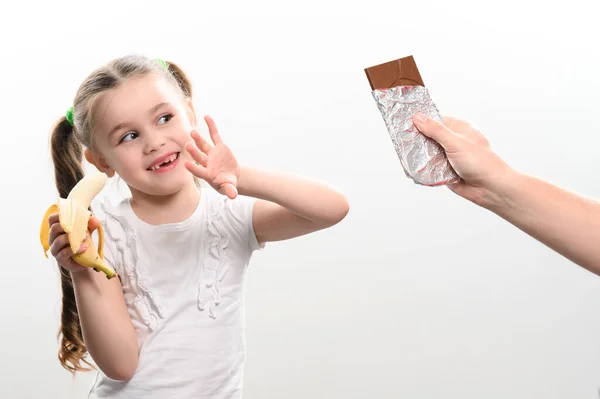 在白色背景下 一个男人的手将巧克力送给手里拿着香蕉的小女孩 复制空间和白色背景 — 图库照片