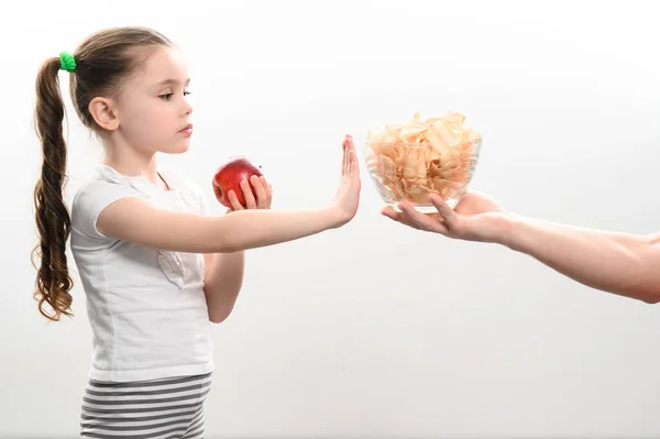A big bowl of chips and apples, a beautiful little girl shows her displeasure with unhealthy food, a child shows a stop hand before the temptation of chips, adults offer chips to a child.