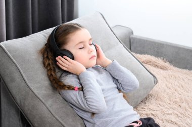 Little girl lies on the sofa and listens to music in headphones and rests, copy space and home rest of the child, children's relaxation with music