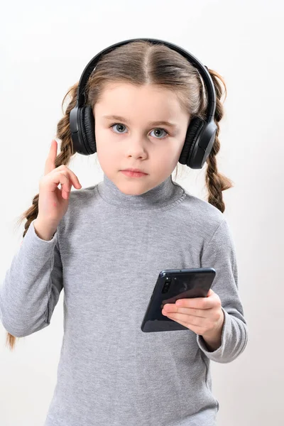 A girl with headphones and a phone on a white background, a girl with pigtails listens to music in headphones, photo with copy space, wireless headphones and a child with a smartphone.