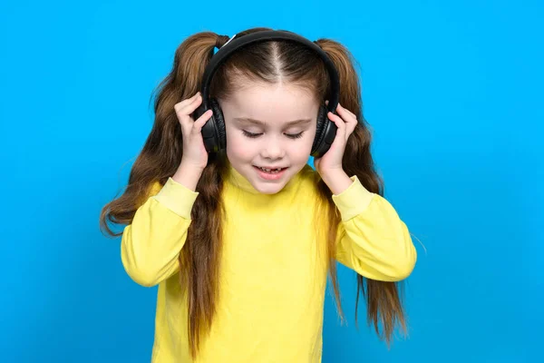 stock image A little girl in a yellow sweater on a blue background listens to music in wireless headphones and holds them in her hands.