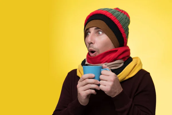 A young man in warm clothes, several hats on a yellow background holds a cup of tea and warms himself during a cold