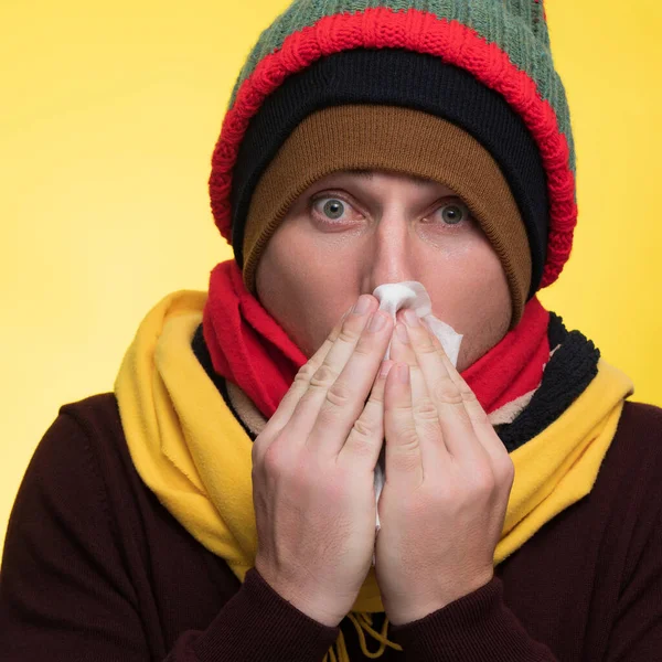 若い悲しい男を身に着けているバーガンディセーター黄色のスカーフ帽子を保持ナプキン組織吹いて鼻のくしゃみ平野黄色の背景スタジオの肖像画健康的なライフスタイル病気の病気の治療寒い季節の概念 — ストック写真
