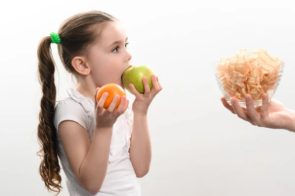 Stock image Big bowl of chips and fruit, beautiful little girl eats an apple, child is given a bowl of chips, white background and copy space, healthy and junk food concept.