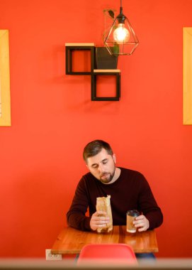 Sakallı bir adam fast food restoranında oturuyor ve elinde shawarma tutuyor ve elinde bir bardak kahve, kırmızı bir duvar ve ahşap bir masa var..