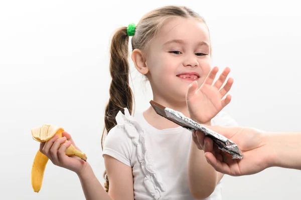 A child is offered chocolate and chooses a banana, on a white background a man\'s hand gives a chocolate to a little girl holding a banana in her hand, copy space and white background.