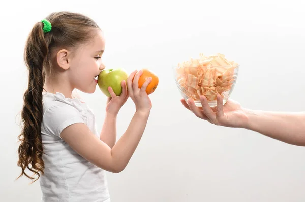 stock image Big bowl of chips and fruit, beautiful little girl eats an apple, child is given a bowl of chips, white background and copy space, healthy and junk food concept.