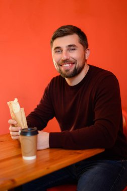 Bir adam bir kafede oturuyor ve gülümsüyor ve elinde bir shawarma tutuyor, fast food restoranı, kırmızı duvar.